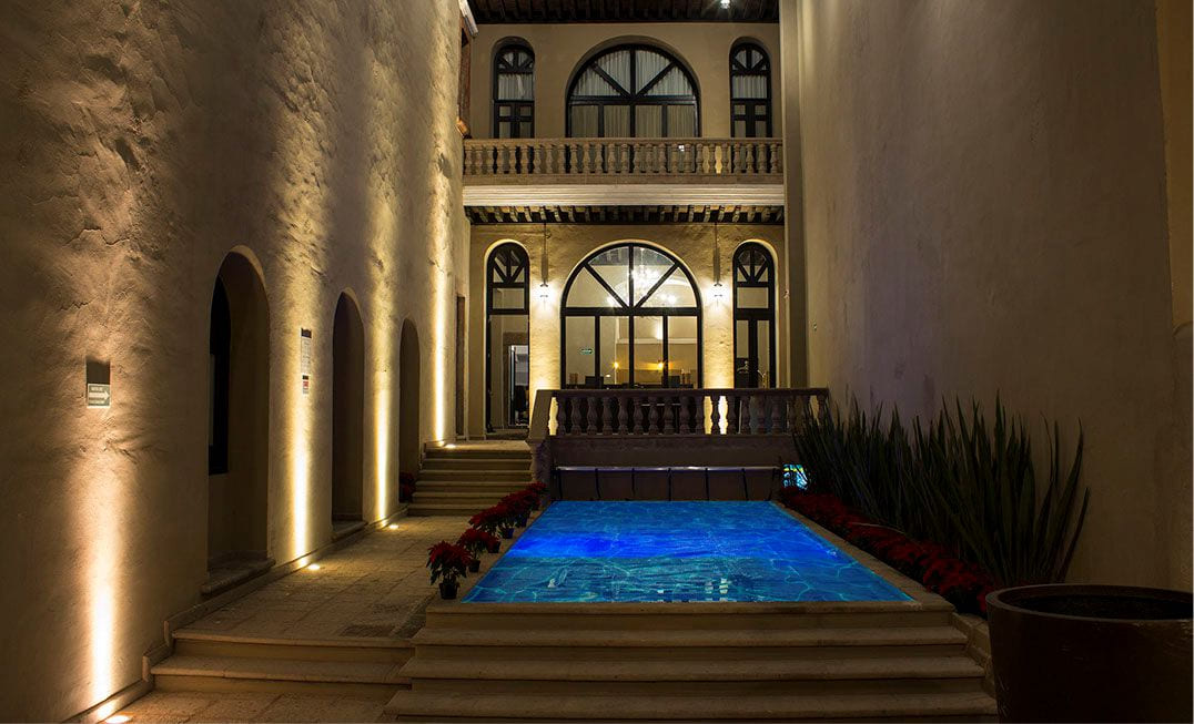 La alberca del hotel iluminada de noche entre los muros de cantera del patio colonial